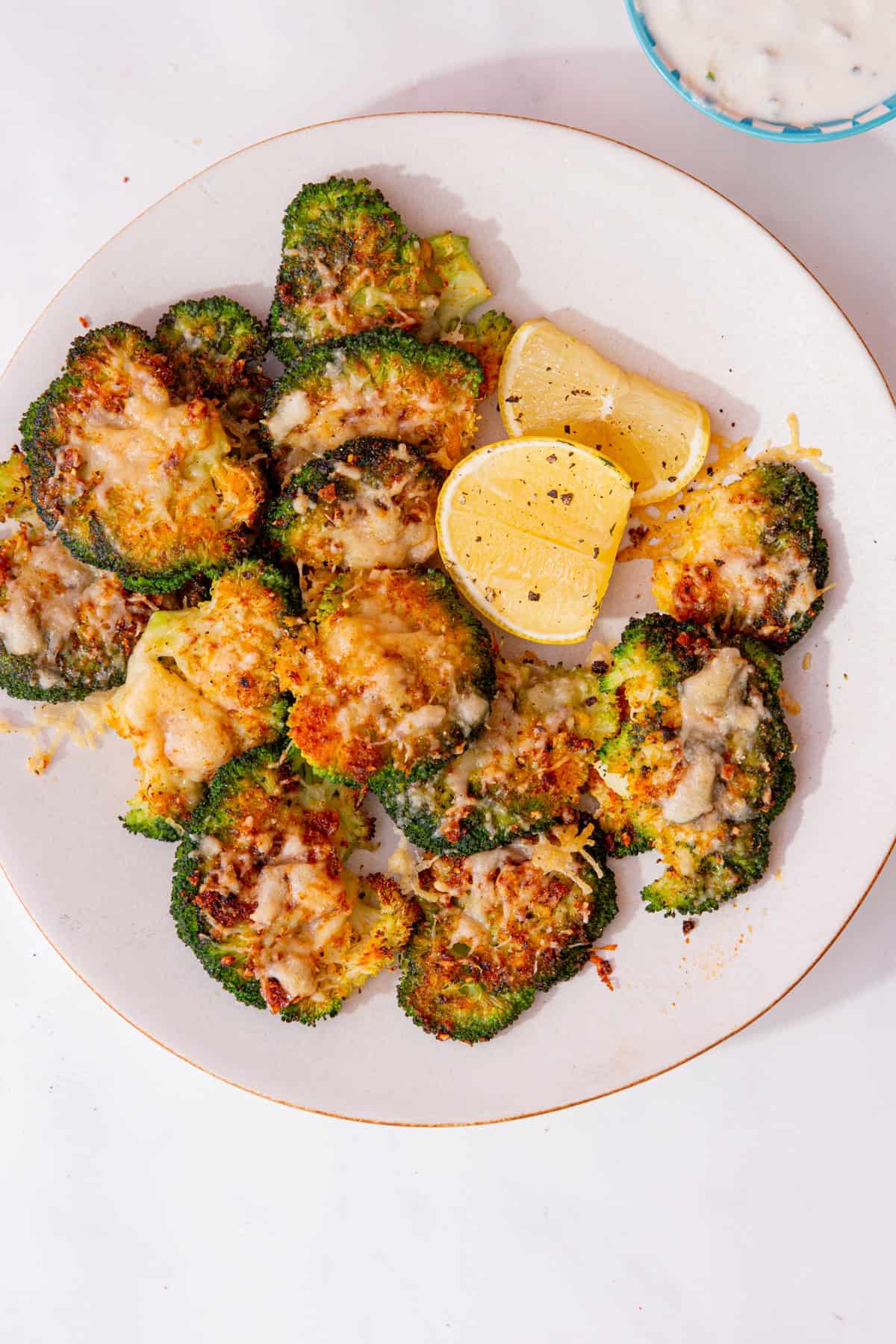 A white plate with roasted broccoli florets with melted cheese and some lemon wedges.