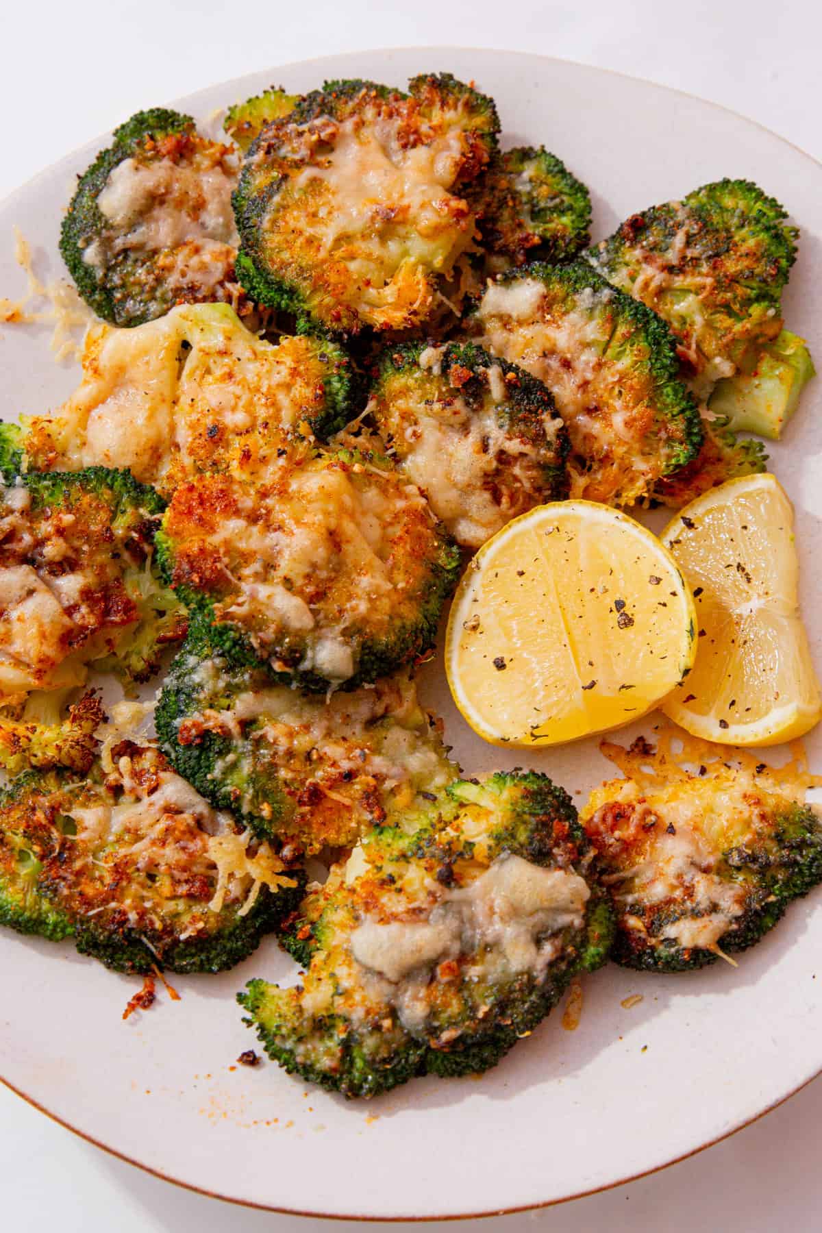 A white plate with roasted broccoli florets with melted cheese covered the golden browned broccoli with a few lemon wedges.