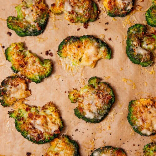 Lots of baked golden browned broccoli florets covered in some cheese scattered over some brown parchment paper.