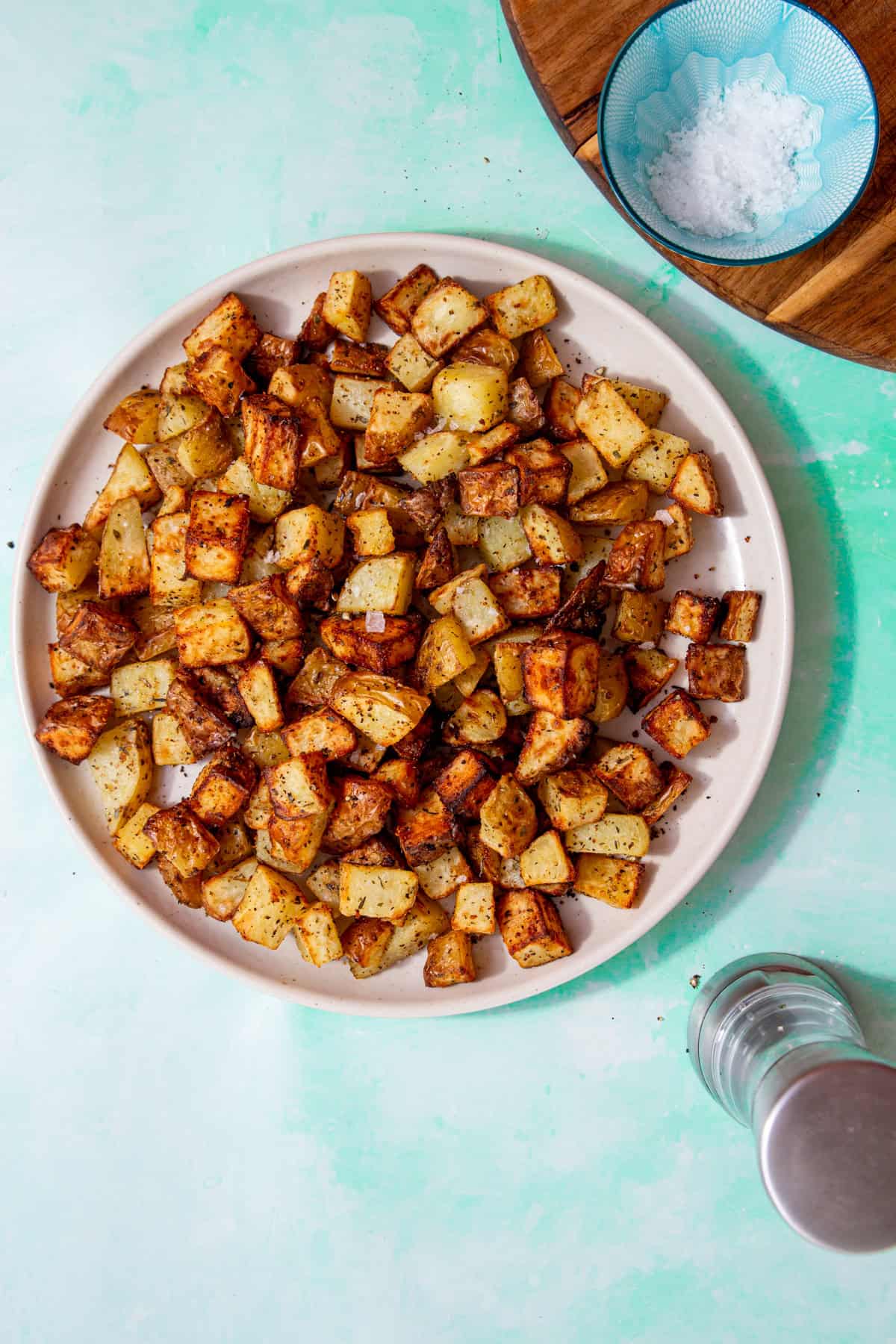 Golden browned diced potatoes with some salt sprinnkled over them on a plate on a pale blue background with a small bowl of salt on a wooden board and a pepper mill below the plate.