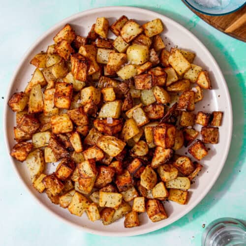 Golden browned diced potatoes with some salt sprinnkled over them on a plate on a pale blue background with a small bowl of salt on a wooden board and a pepper mill next to the plate.