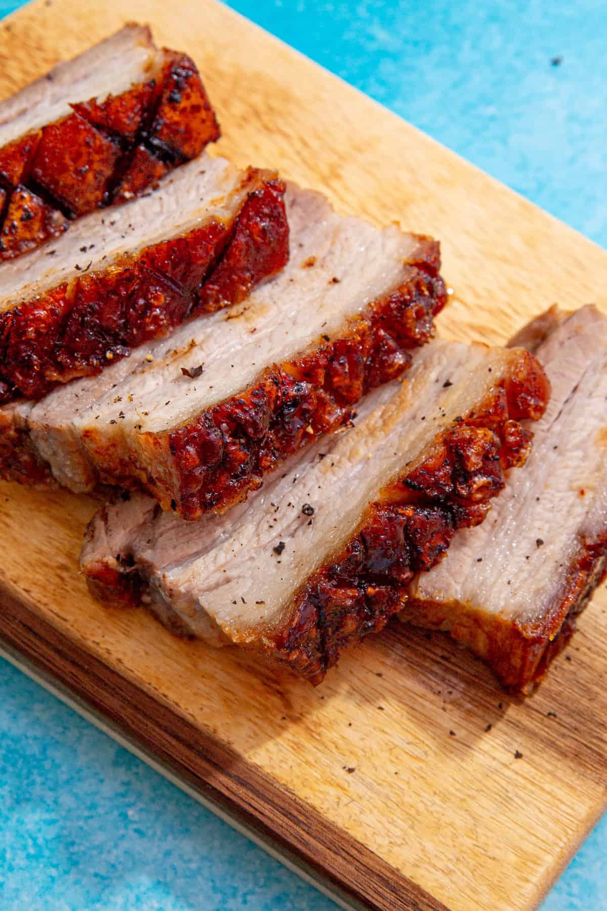 5 slices of pork belly with a golden browned crackling topping on a wooden chopping board on a blue background.