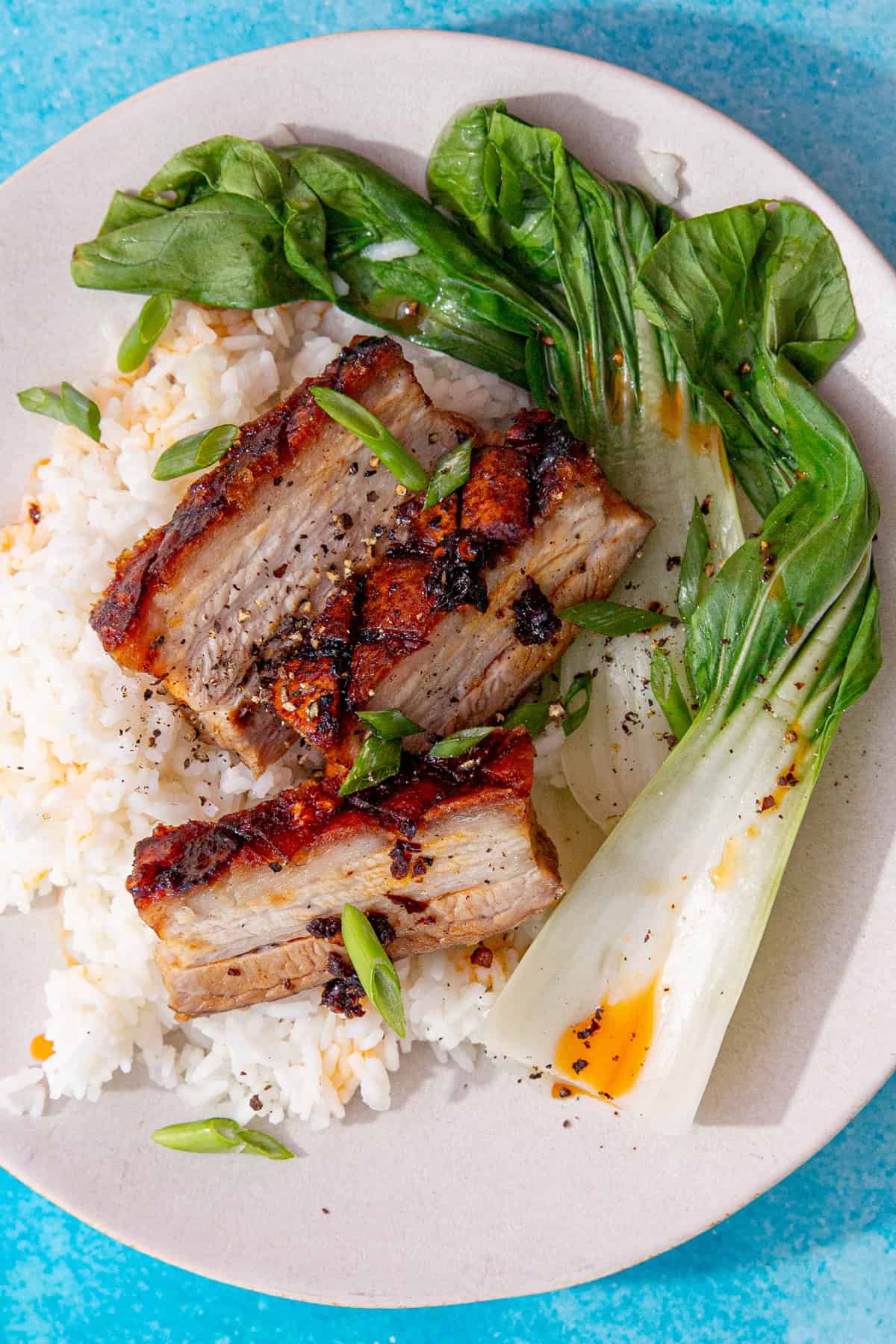 A plate with 3 thick slices of pork belly with a golden browned crackling on a bed of rice with some pak choi.