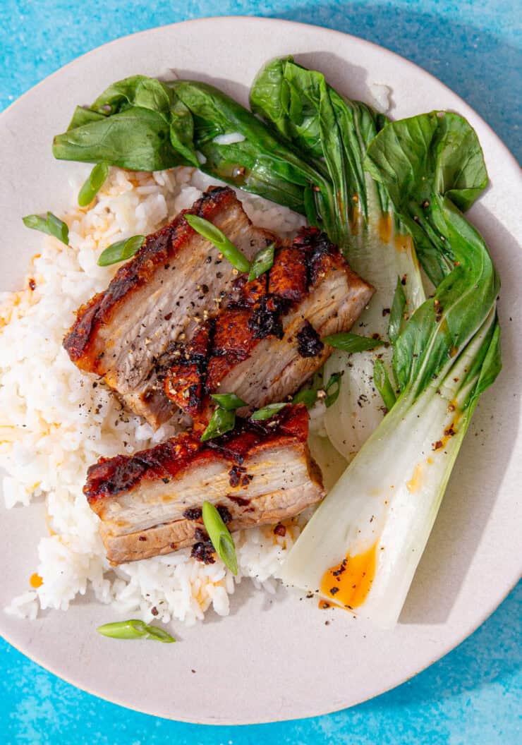 3 slices of golden bowned pork on a bed of rice with pac choi on a blue background.