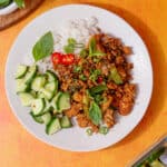 Chicken mince stir fry recipe on a plate with sliced cucumber, rice, Thai basil, spring onions and chillies on a plate.