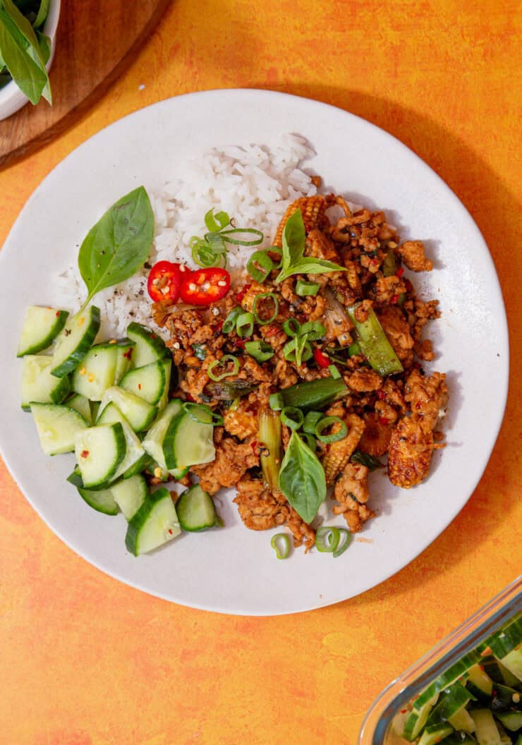 Chicken mince stir fry recipe on a plate with sliced cucumber, rice, Thai basil, spring onions and chillies on a plate.