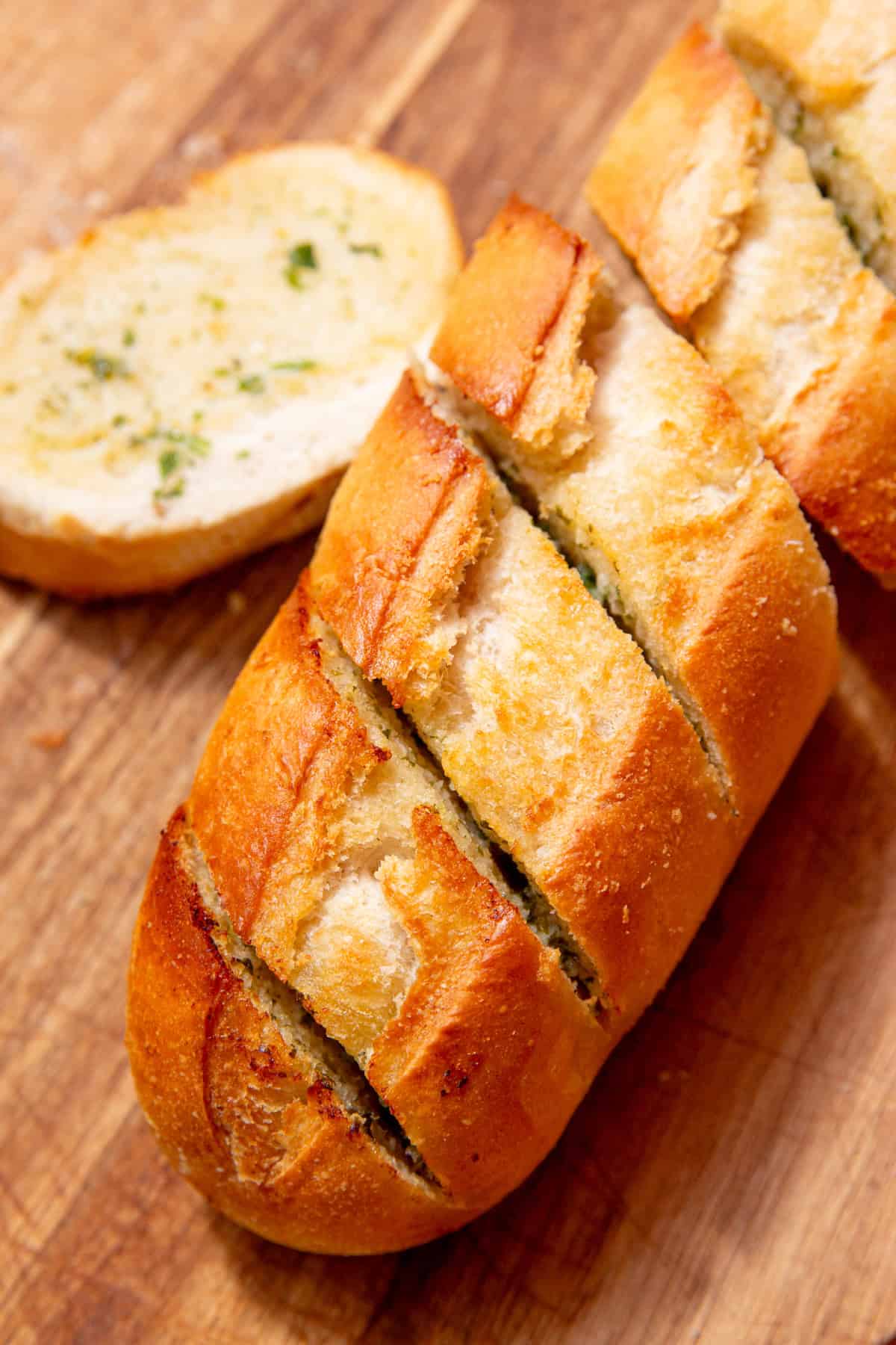 A golden browned partly sliced garlic bread baguette with a separate piece next to the baguette all on a chopping board.