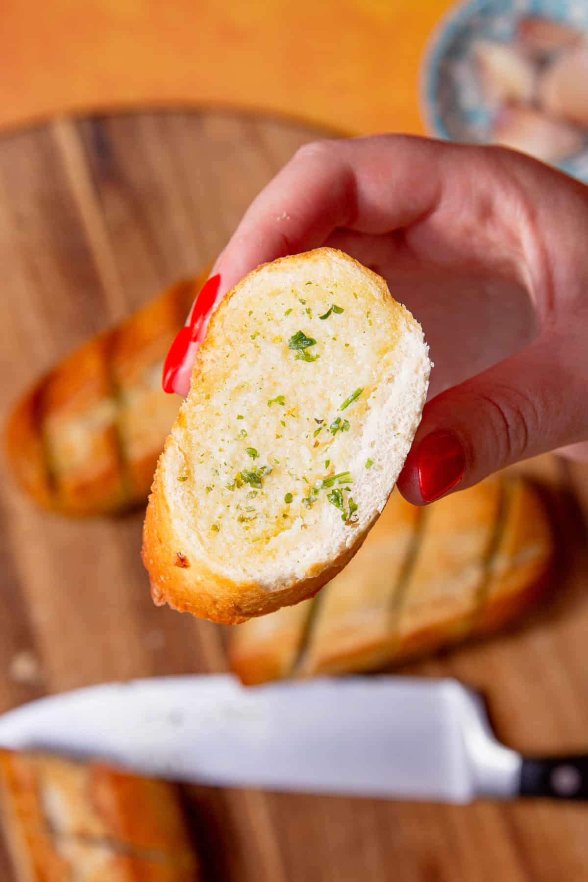 A hand holding a piece of garlic bread with a garlic bread baguette faded in background on a wooden board.