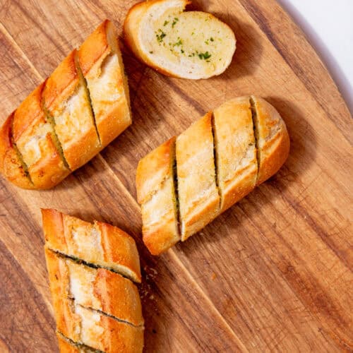 A golden browned, baked garlic bread baguette broken into 4 pieces with pre slice marks on a wooden chopping board.