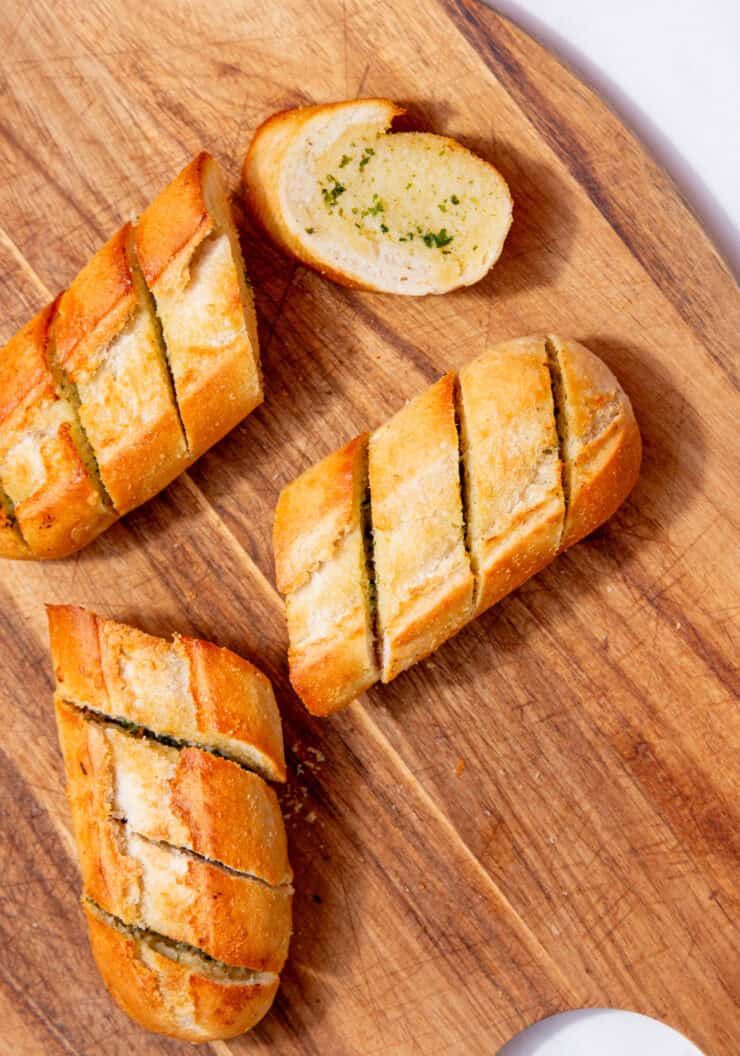 A golden browned, baked garlic bread baguette broken into 4 pieces with pre slice marks on a wooden chopping board.