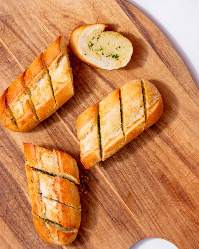 A golden browned, baked garlic bread baguette broken into 4 pieces with pre slice marks on a wooden chopping board.