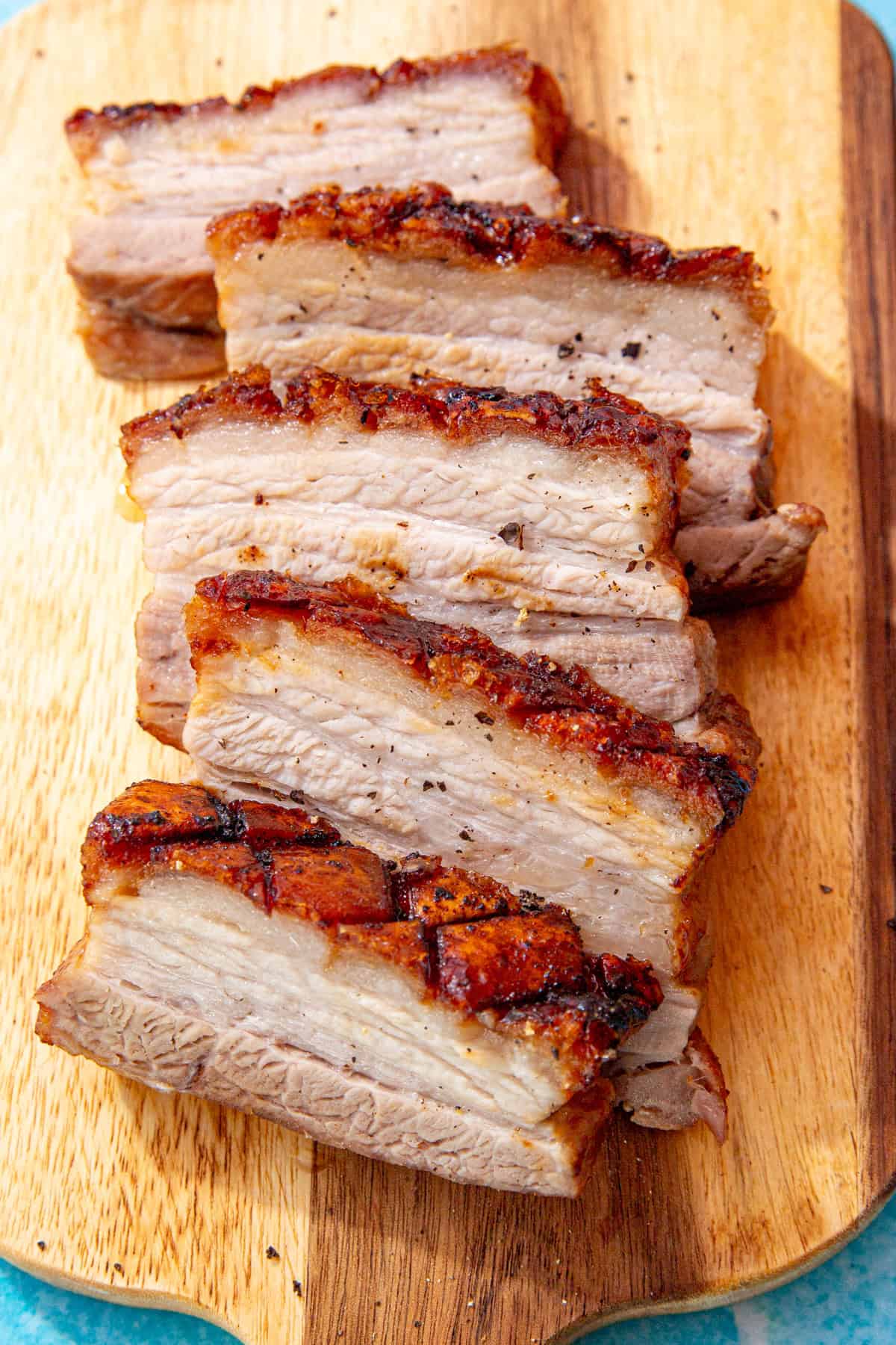 5 slices of pork belly with a golden browned crackling topping on a wooden chopping board.