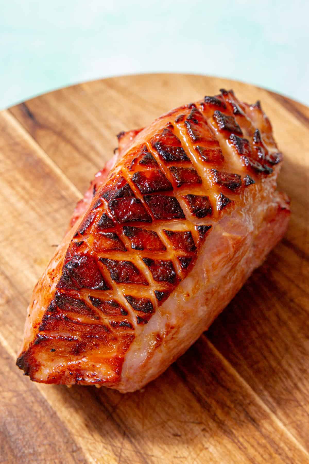 A large piece of gammon with golden browned, scored and glazed skin laying on a wooden chopping board.