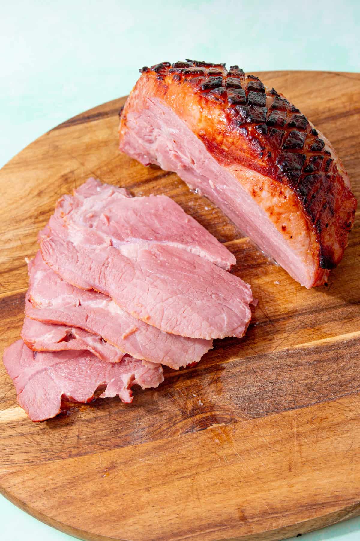 A large piece of gammon with golden browned, scored and glazed skin  and lots of slices of gammon on a wooden chopping board.