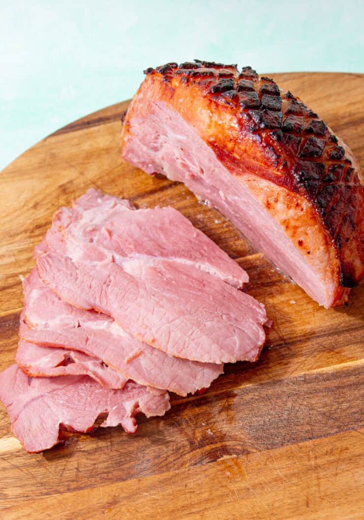 A large piece of gammon with golden browned, scored and glazed skin woth around 4 slices of gammon next to the joint on a wooden chopping board.