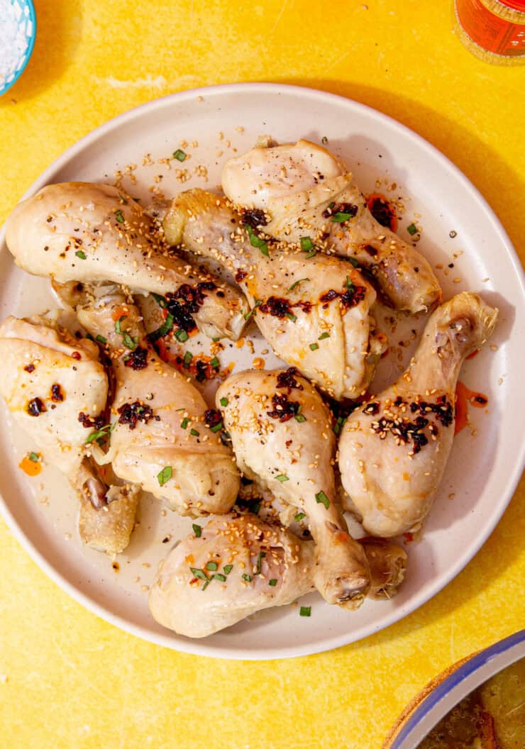 Boiled chicken drumsticks on a plate topped with chilli oil, sesame seeds and chopped basil on a yellow background with a small bowl of salt, a jar of spices and the saucepan in partial view below.