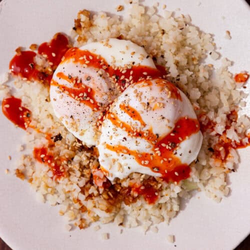 2 cooked poached eggs on a bed of rice with a drizzle of sriracha over the top on a white plate.