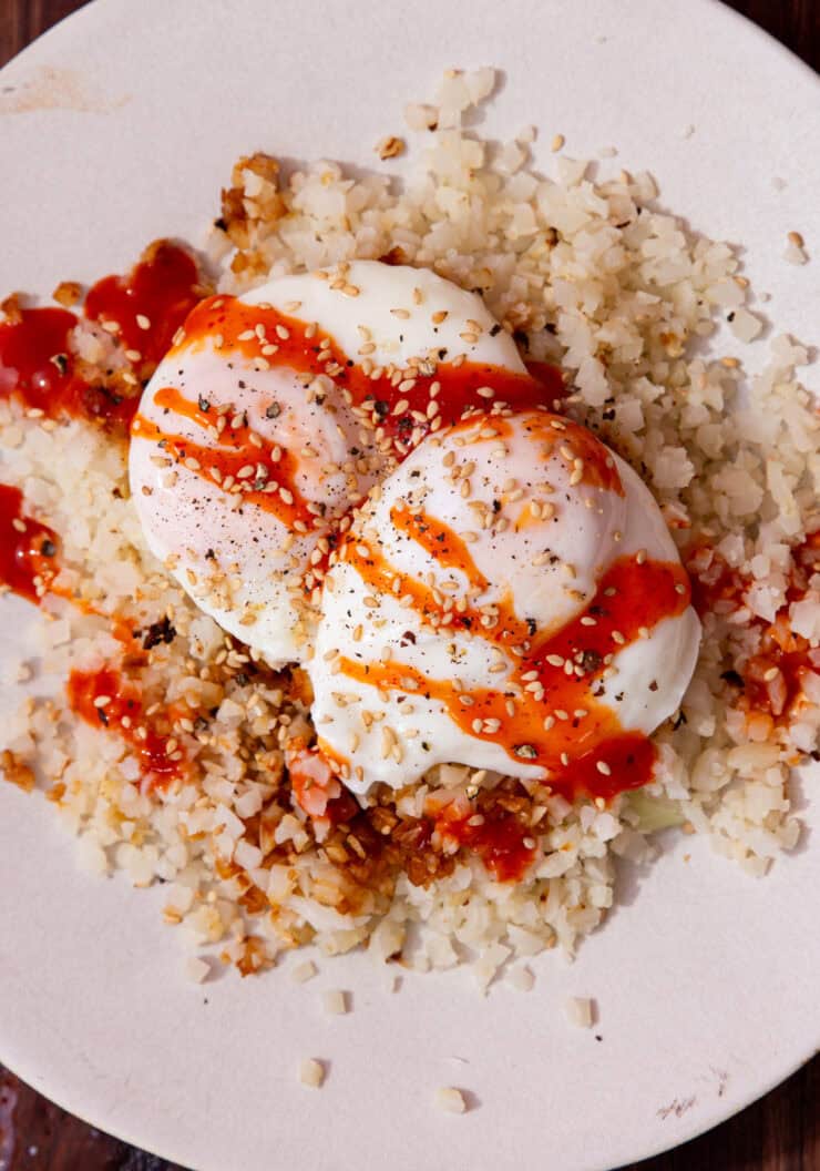 2 cooked poached eggs on a bed of rice with a drizzle of sriracha over the top on a white plate.