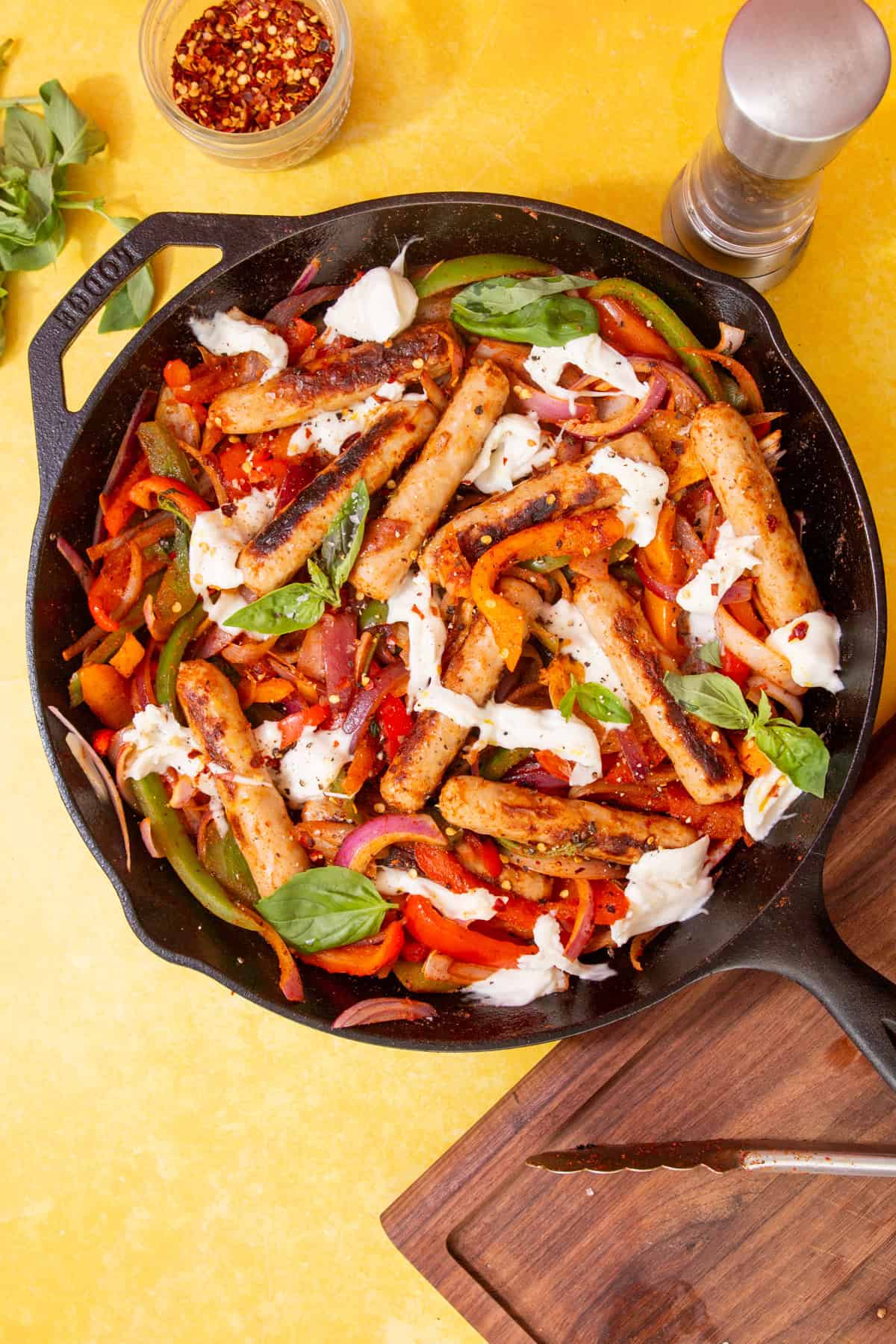 Golden browned sausages, peppers, onion topped with moczarella cheese topped with fresh basil in a skillet pan on a yellow background with chillies flakes, fresh basil and a pepper pot in view.