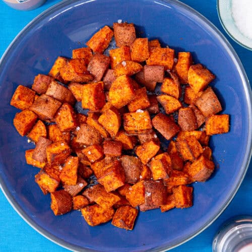 Cooked and browned sweet potato cubes on a blue plate on a blue background next to some salt in a bowl and a container with spices.