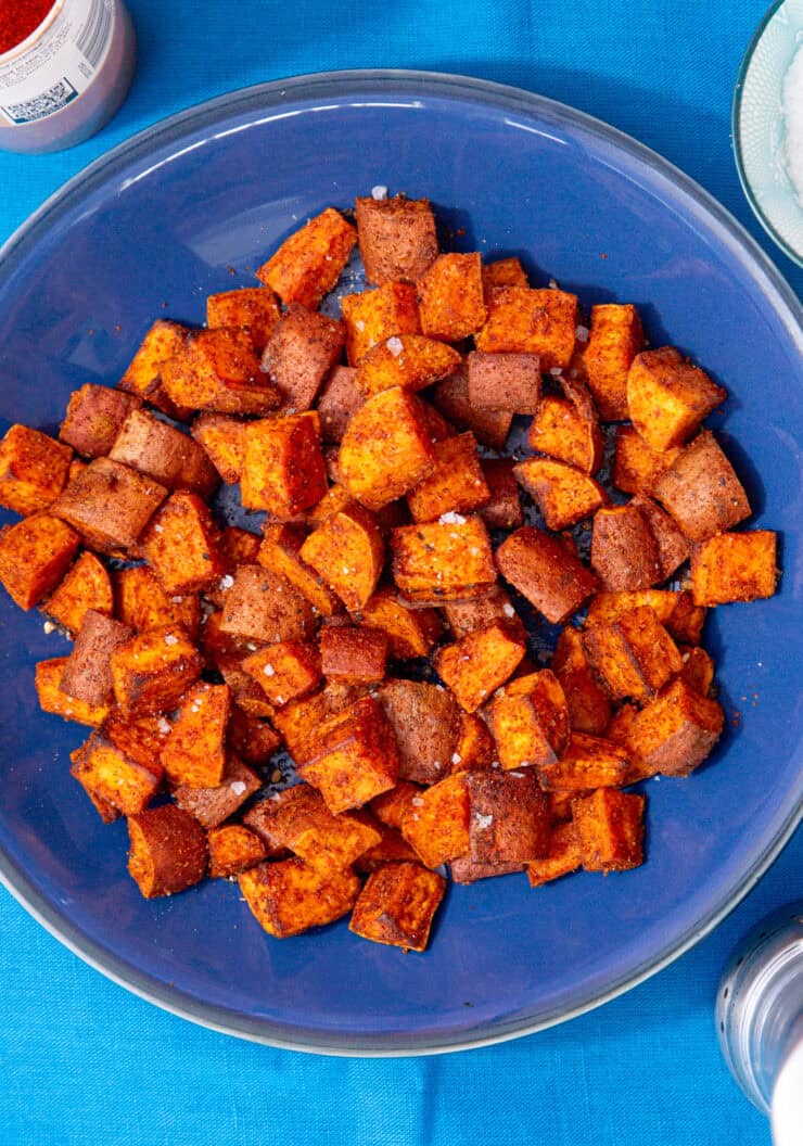 Cooked and browned sweet potato cubes on a blue plate on a blue background next to some salt in a bowl and a container with spices.