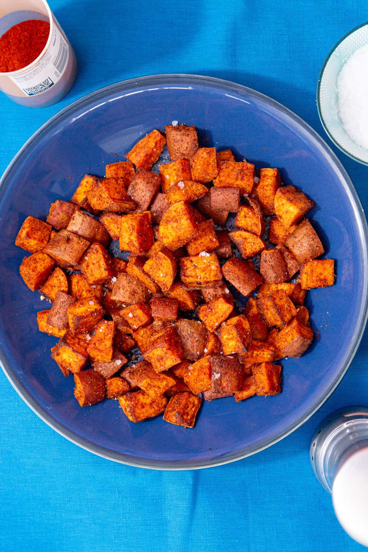 Lots of cooked and browned sweet potato cubes on a blue plate on a blue background next to some salt in a bowl and a container with spices.