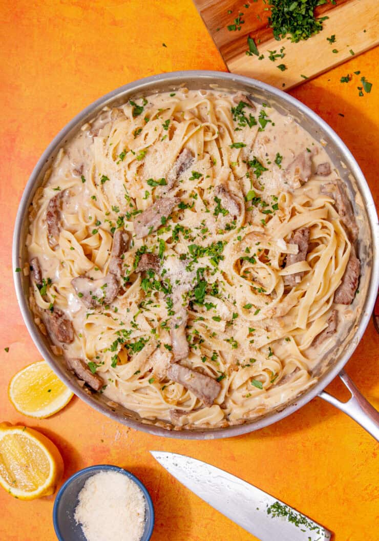 Tagliatella in a creamy sauce with steak strips and some green herbs in a large pan on an orange background with lemon wedges scattered around.