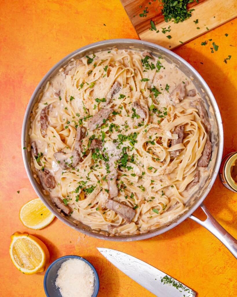 Tagliatella in a creamy sauce with steak strips and some green herbs in a large pan on an orange background with lemon wedges scattered around.