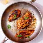 A large stainless steel frying panwith 3 pieces of seared chicken breast with fresh green herbs.