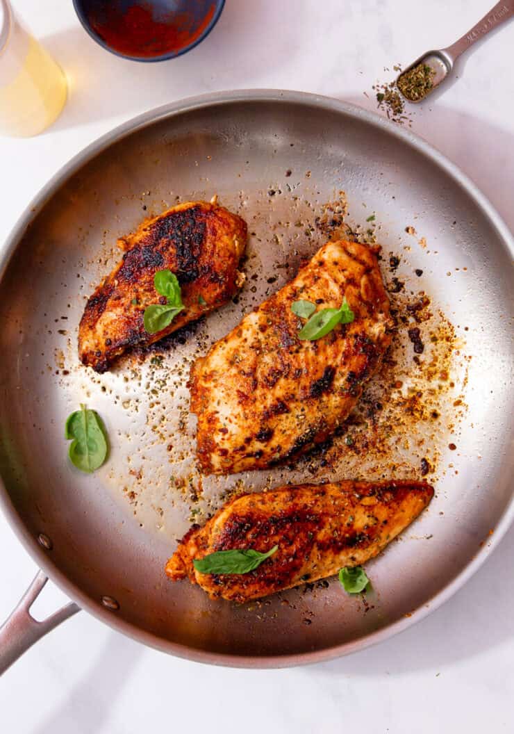 A large stainless steel frying panwith 3 pieces of seared chicken breast with fresh green herbs.