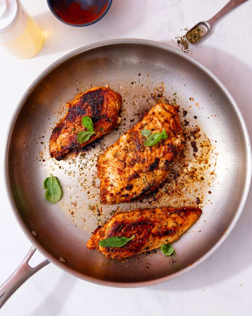 A large stainless steel frying panwith 3 pieces of seared chicken breast with fresh green herbs.