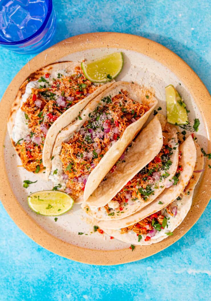 4 chicken tacos with other toppings laid out on a plate with some lime wedges on a blue background with a glass of water with ice.