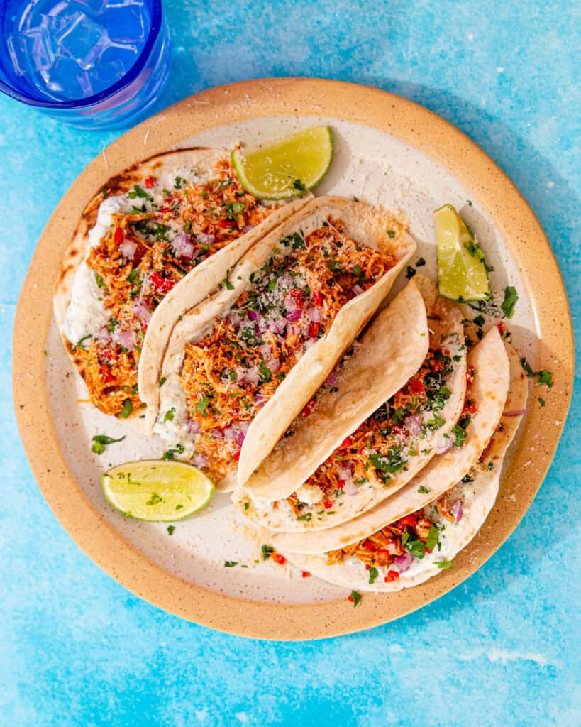 4 chicken tacos with other toppings laid out on a plate with some lime wedges on a blue background with a glass of water with ice.