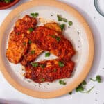 4 pieces of pork covered in a reddish sauce, some seasoning and some fresh parsley sprigs on a plate on a white background.