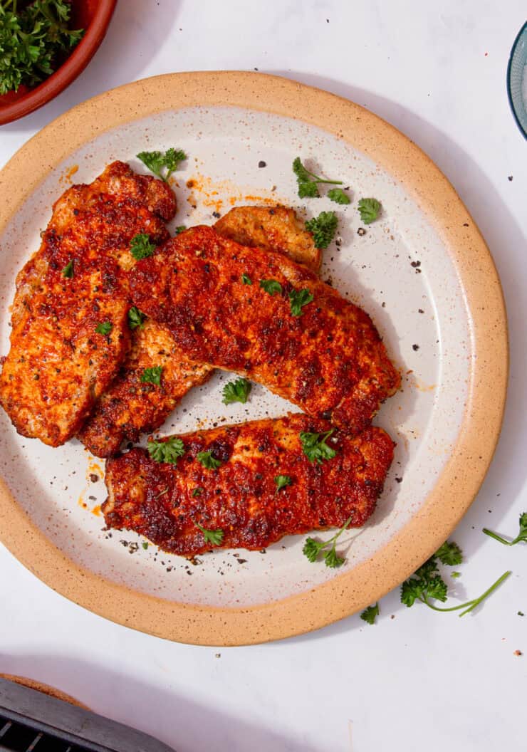 4 pieces of pork covered in a reddish sauce, some seasoning and some fresh parsley sprigs on a plate on a white background.