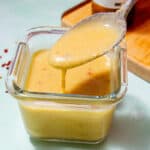 A square, glass container with parmesan vinaigrette dressing with a spoonful of dressing being poured, next a chopping board.