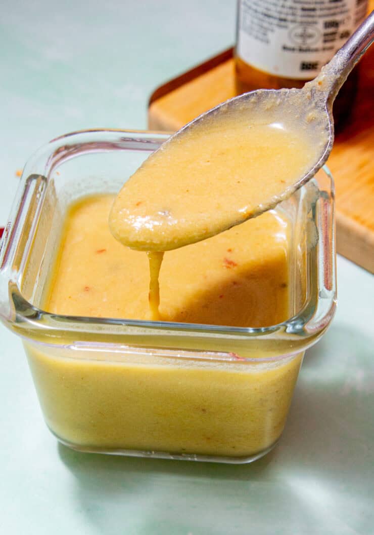 A square, glass container with parmesan vinaigrette dressing with a spoonful of dressing being poured, next a chopping board.