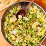 A stainless steel frying pan with chopped savoy cabbage leaves and some pistatio nuts, stirred with a wooden spoon next to a chopping board with lemon wedges in partial view.
