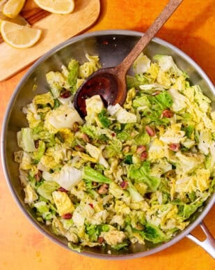 A stainless steel frying pan with chopped savoy cabbage leaves and some pistatio nuts, stirred with a wooden spoon next to a chopping board with lemon wedges in partial view.