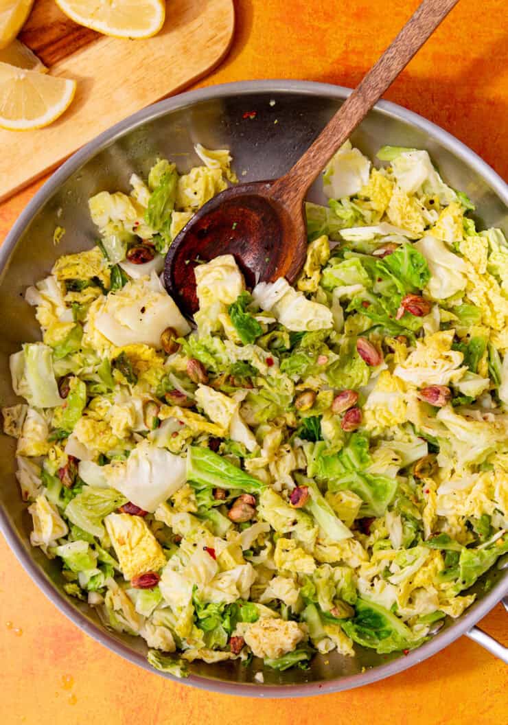 A stainless steel frying pan with chopped savoy cabbage leaves and some pistatio nuts, stirred with a wooden spoon next to a chopping board with lemon wedges in partial view.