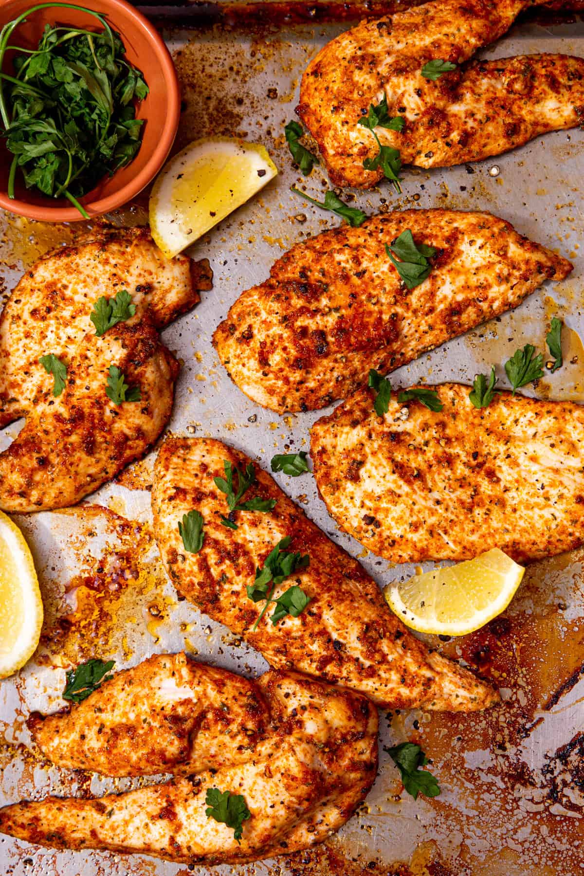 Thin slices of cooked and seasoned chicken on a baking tray with lemon wedges and fresh herbs.