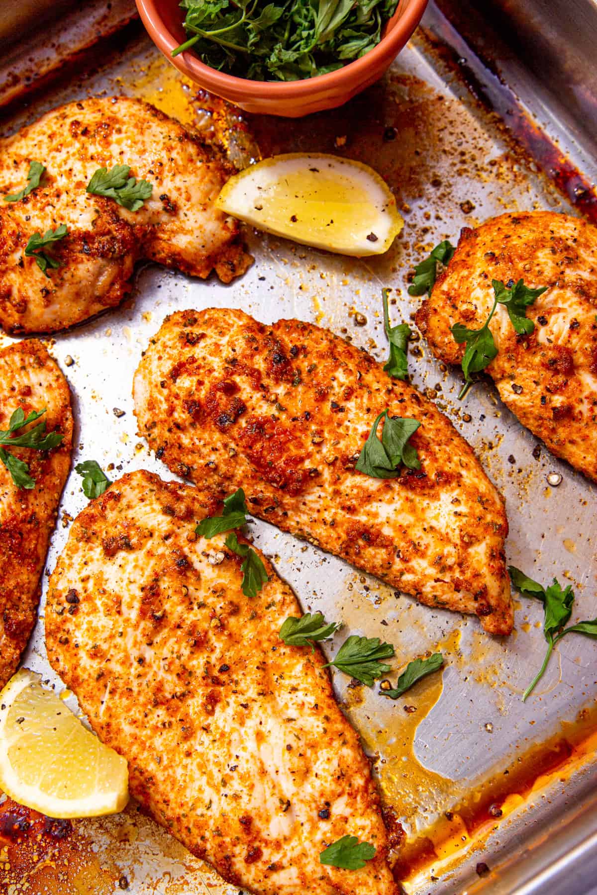 Thin slices of cooked and seasoned chicken on a baking tray with lemon wedges and fresh parsley in a bowl on the baking tray.