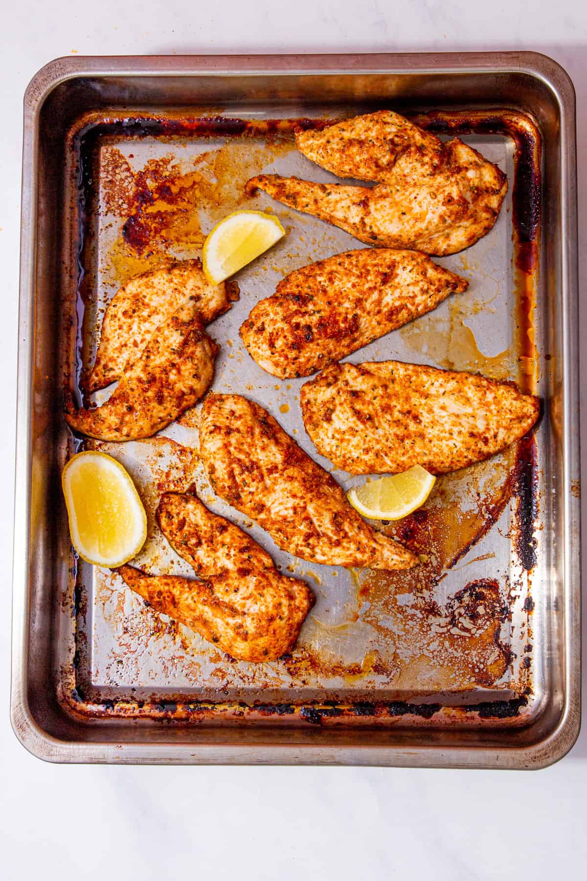 Thin slices of cooked and seasoned chicken on a baking tray with lemon wedges.