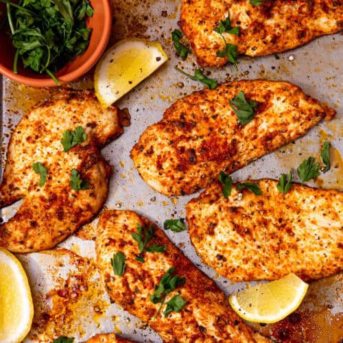 Thin, seasoned, cooked chicken breast fillets on a baking tray with some lemon wedges and fresh parsley.