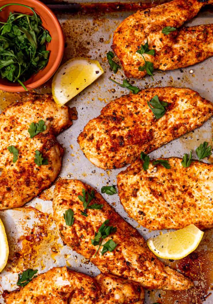 Thin, seasoned, cooked chicken breast fillets on a baking tray with some lemon wedges and fresh parsley.