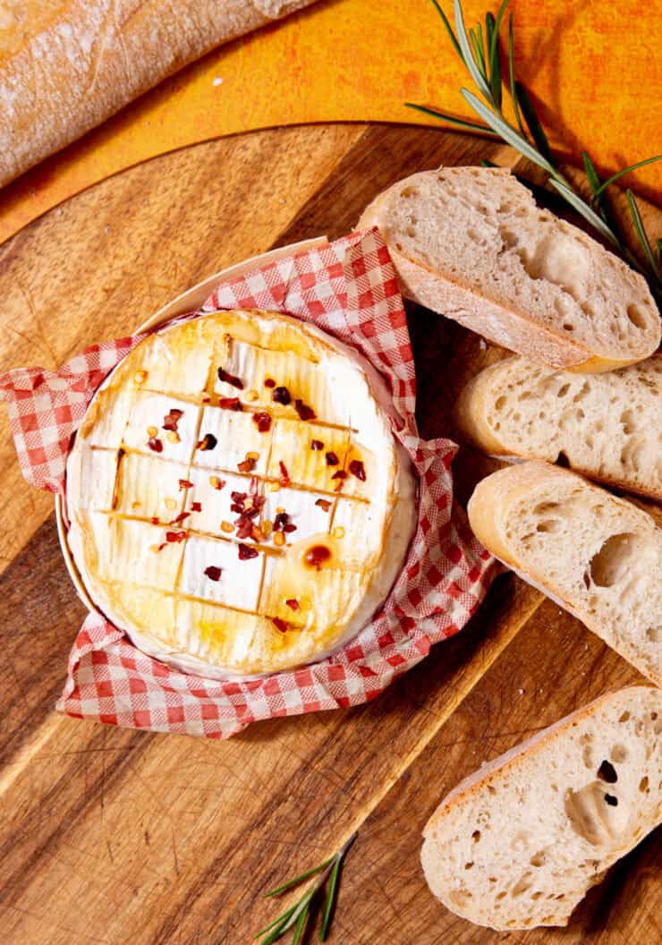 image of baked camembert on a red checkered napkin on a cutting board next to sliced bread and a sprig of rosemary