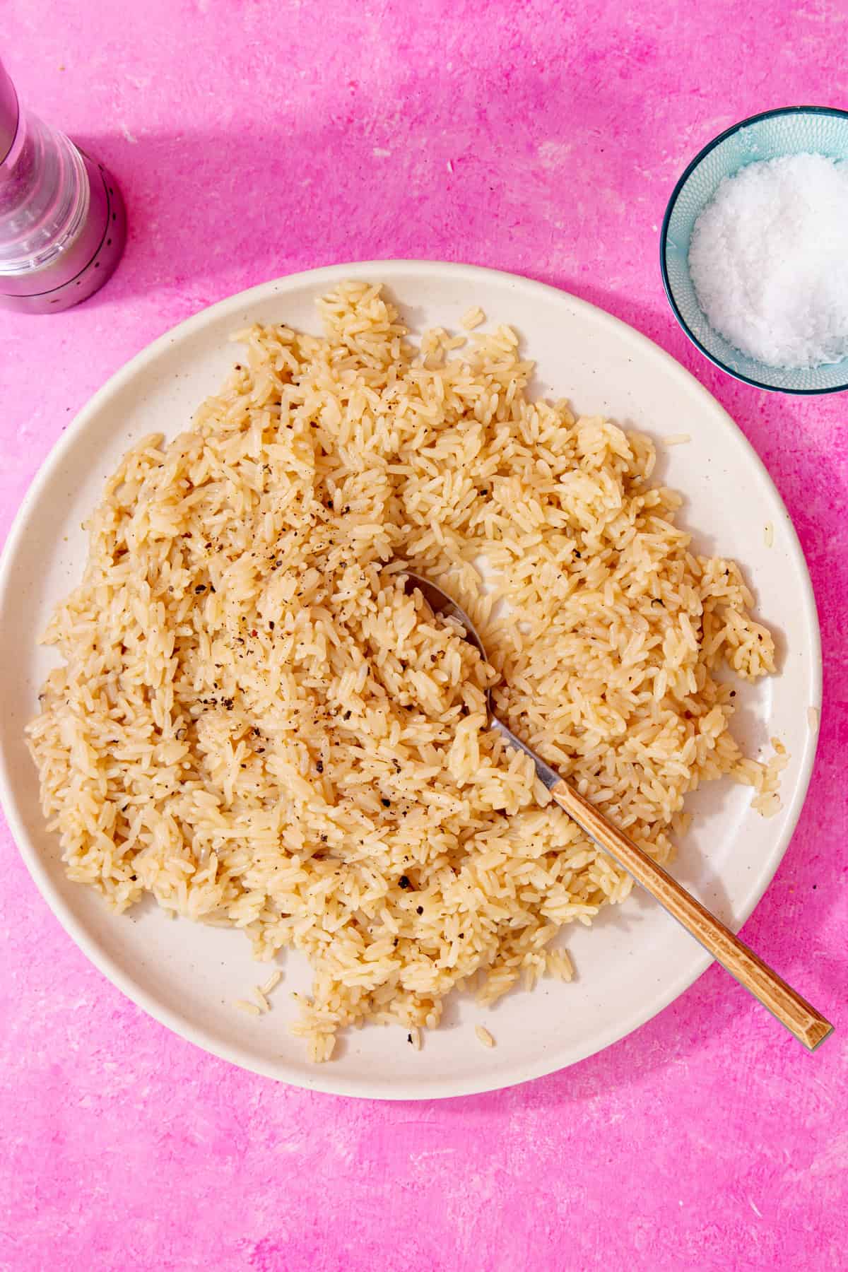 Creamy buttered rice with chicken stock  and sprinkled with pepper on a white plate with a spoon all on a pink background .