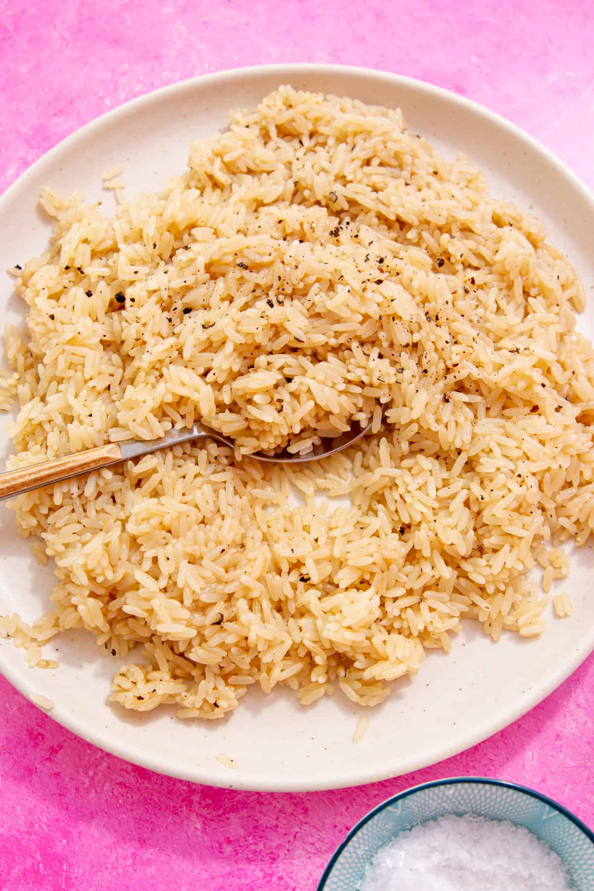 Close up of fluffy rice with chicken broth sprinkled with ground pepper and a spoon taking a scoop.  All on a white plate on a pink background.