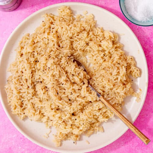 perfectly cooked and seasoned rice with chicken broth and butter on a white plate with a spoon in it all on a pink background.