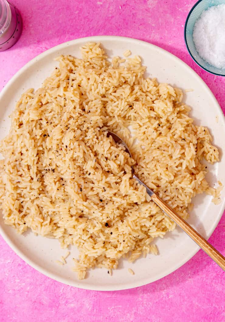 perfectly cooked and seasoned rice with chicken broth and butter on a white plate with a spoon in it all on a pink background.